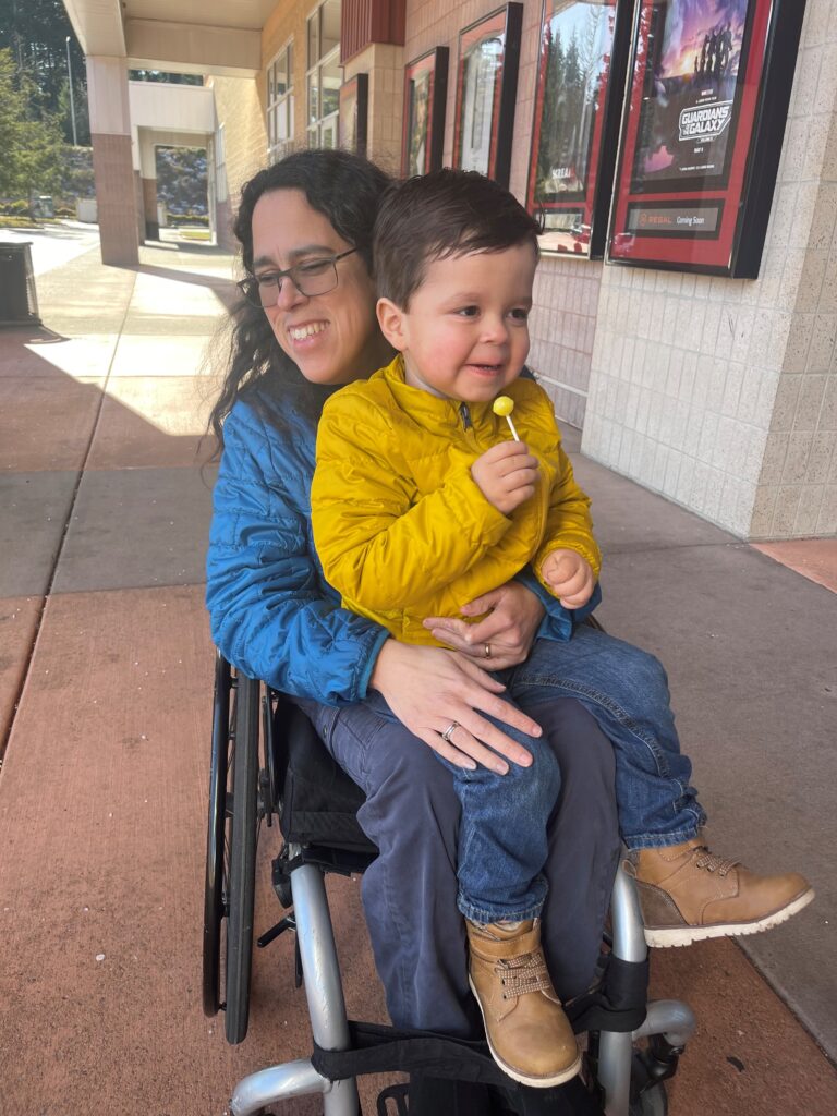 Photo of Jessie Carver wearing a blue jacket and pants sitting in a wheelchair with son sitting on lap wearing a yellow jacket, jeans, and brown boots
