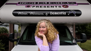 Image of a woman with median length curly blonde hair in front of a white van that reads Women, Wine, and Dementia